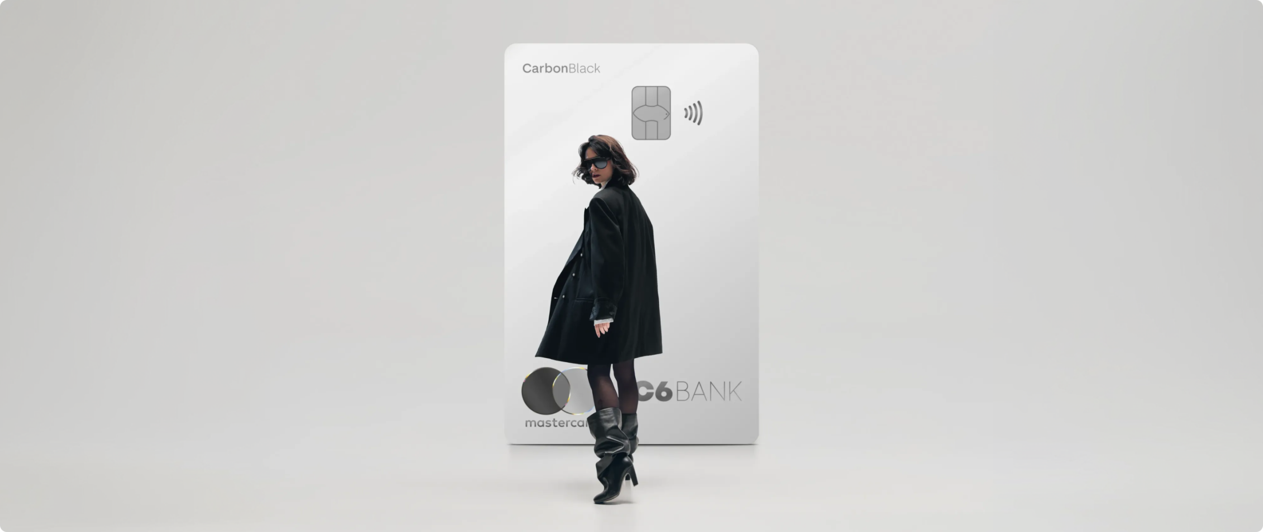 Mulher em frente a cartão Mulher de preto caminha à frente de um Cartão C6 Carbon Mastercard Black prata em tamanho ampliado, sobre fundo claro.